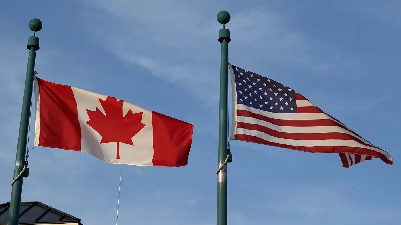 Canadian and American flags flying side by side on tall flagpoles against a clear blue sky. The Canadian flag, with a red maple leaf, is on the left, and the United States flag, with stars and stripes, is on the right. The flags are gently waving in the wind.