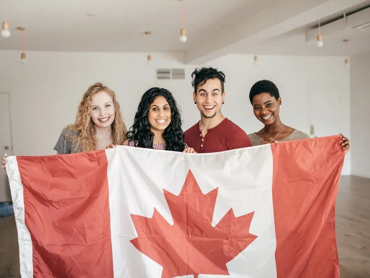 Four smiling young adults standing indoors and proudly holding a large Canadian flag. The group is diverse, with two women on either side and one man and one woman in the middle, all looking at the camera in a well-lit room with minimal decor.