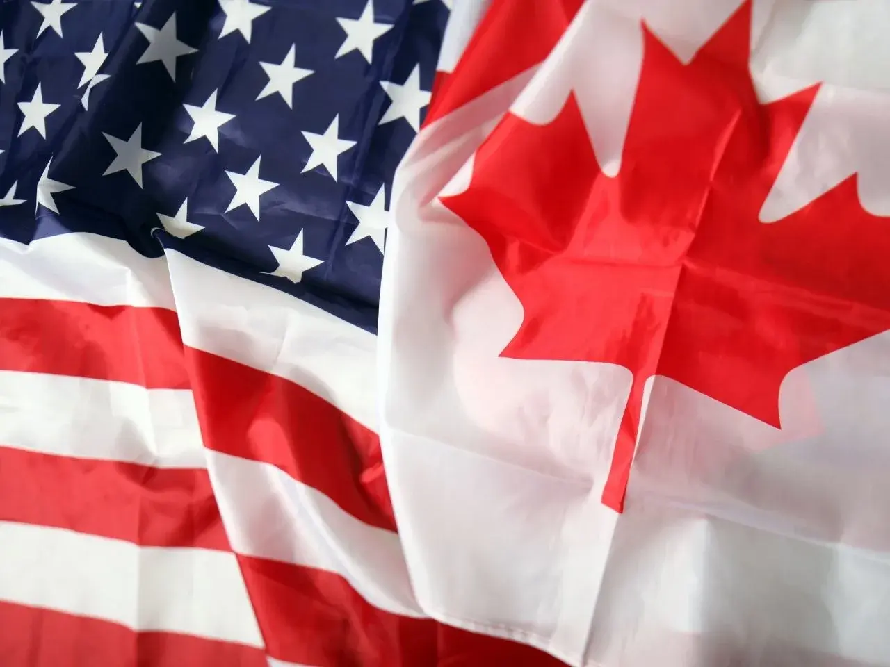 Close-up of overlapping United States and Canadian flags, showing the red maple leaf of Canada and the stars and stripes of the U.S. flag. The fabric appears slightly wrinkled, emphasizing texture and contrast between the two national symbols.