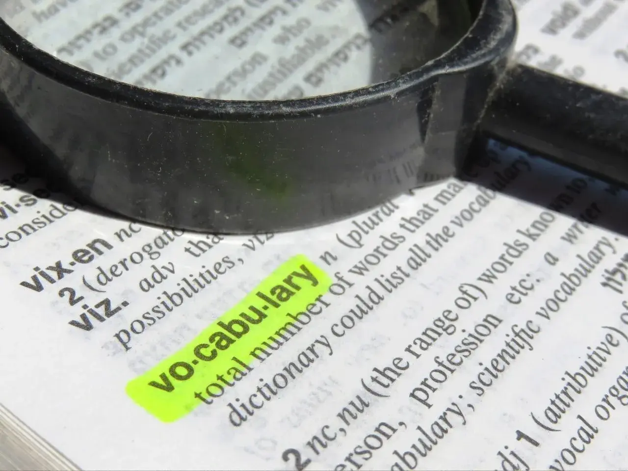 Close-up of a dictionary page with the word “vocabulary” highlighted in bright yellow. A black magnifying glass lies on top, partially enlarging the surrounding text and emphasizing the word’s definition.