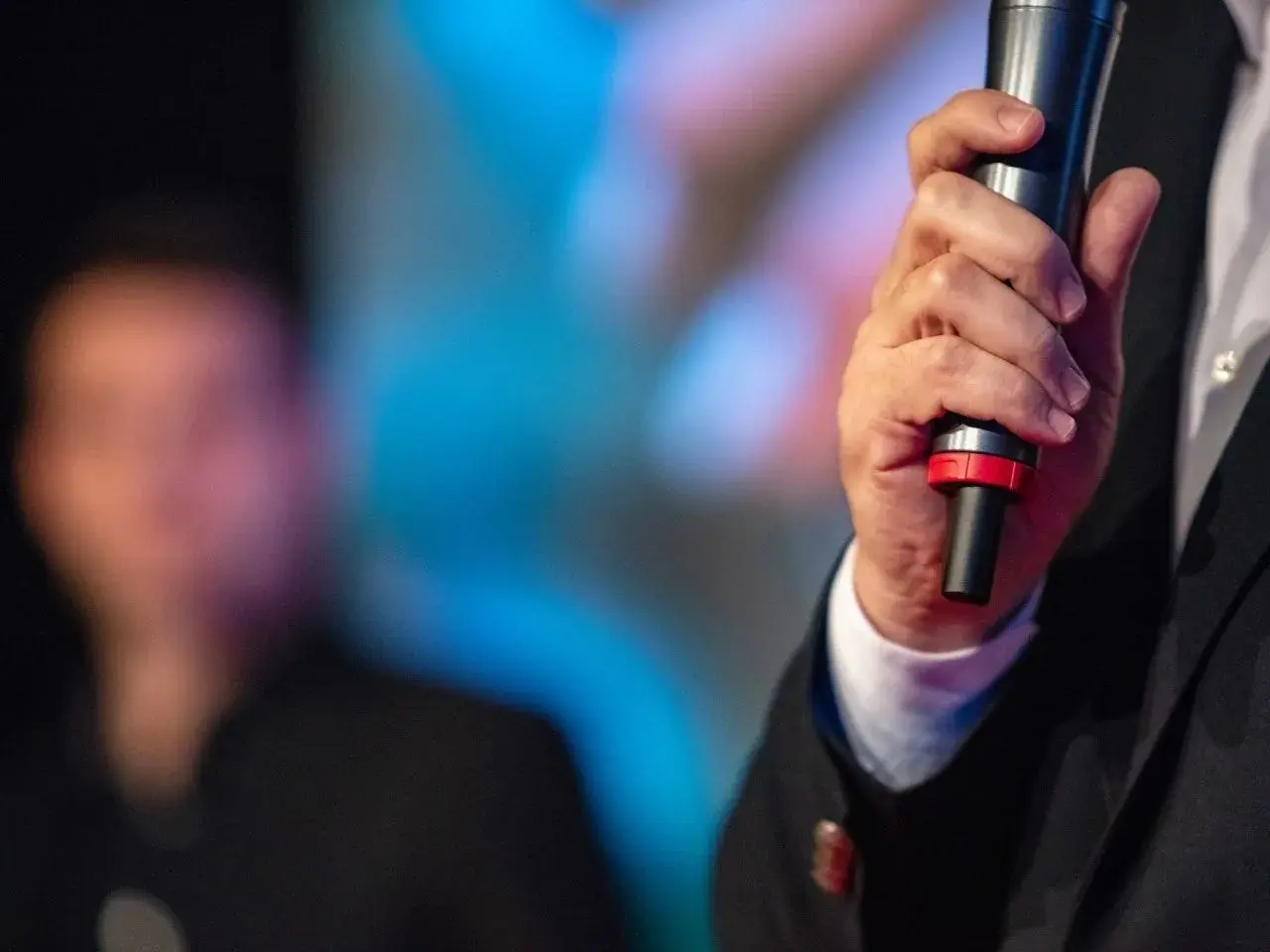 Close-up of a person holding a microphone, dressed in a suit, likely preparing to speak or give a presentation. The background is blurred with colourful stage lighting and an indistinct figure in the distance, suggesting a formal or public speaking event.