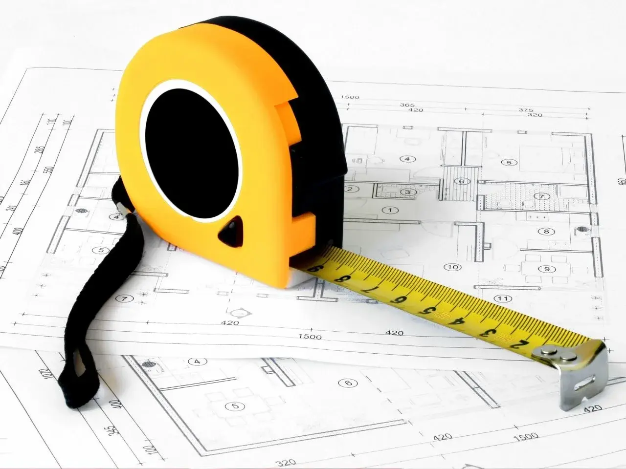 A yellow and black tape measure partially extended, resting on a detailed architectural blueprint. The blueprint contains room layouts and measurements, indicating a construction or design project in progress.