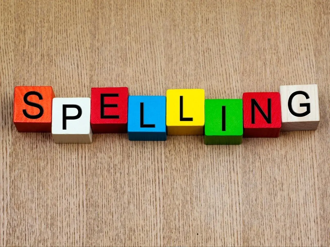 Colourful wooden blocks arranged on a wooden surface to spell the word “SPELLING,” with each letter printed in black on a different coloured block.
