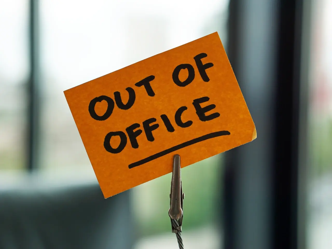A handwritten orange sticky note clipped upright, with the words “OUT OF OFFICE” in bold black letters, set against a blurred office background.