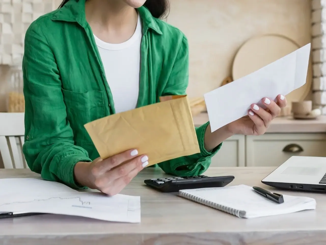 A woman in a green shirt sitting at a desk, holding a padded envelope in one hand and a white letter envelope in the other, surrounded by a calculator, laptop, and documents.