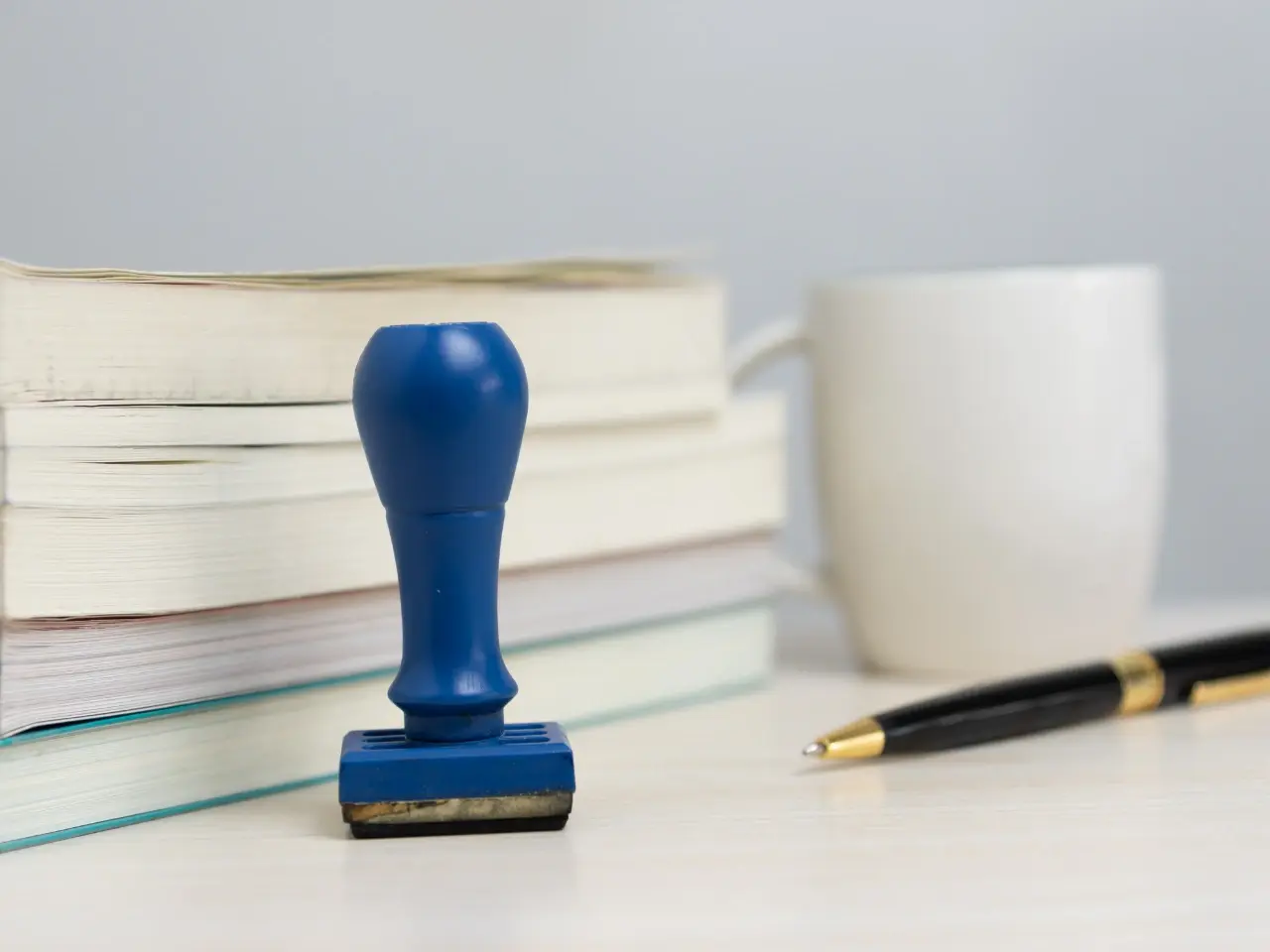 A blue rubber stamp standing upright on a desk beside a stack of books, a white coffee mug, and a black-and-gold pen.