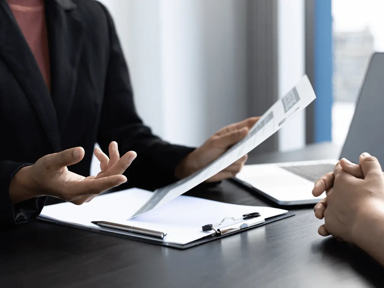 A legal professional in a black suit discussing documents during a meeting, holding a form while explaining requirements to a client.