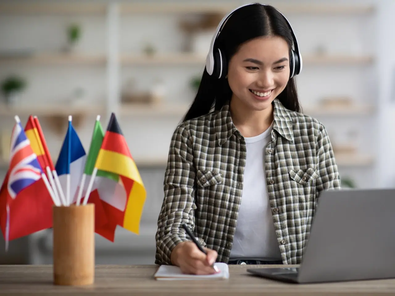 Smiling translator with headphones taking notes during an online session, with multiple international flags on the desk, representing multilingual certified translation services.