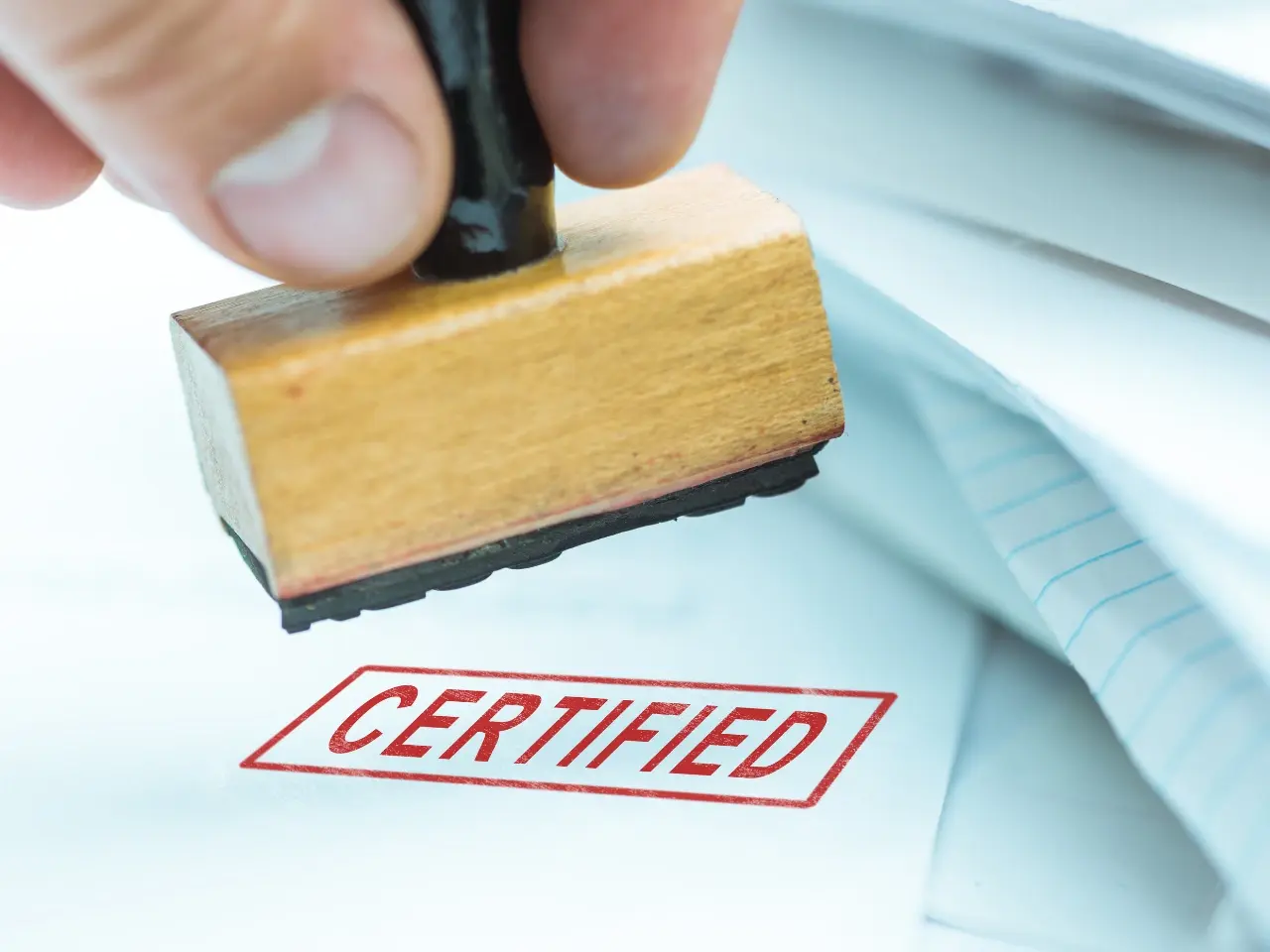 Close-up of a wooden stamp marking the word “CERTIFIED” in red on a document, representing the official validation of translated papers.