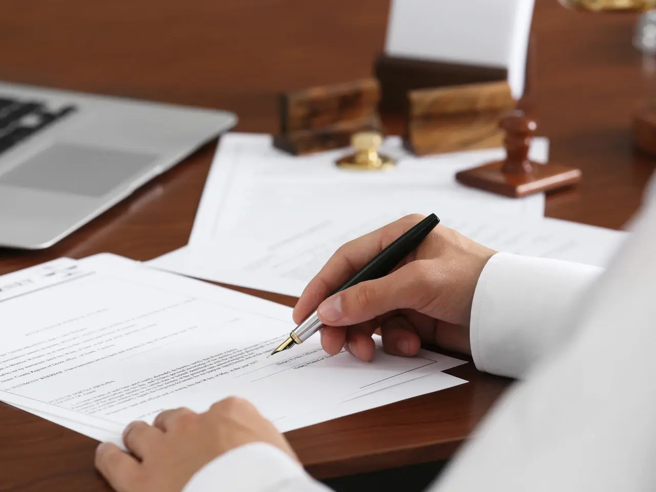 Close-up of a lawyer signing legal documents with a fountain pen, with official stamps and a laptop on the desk in the background.