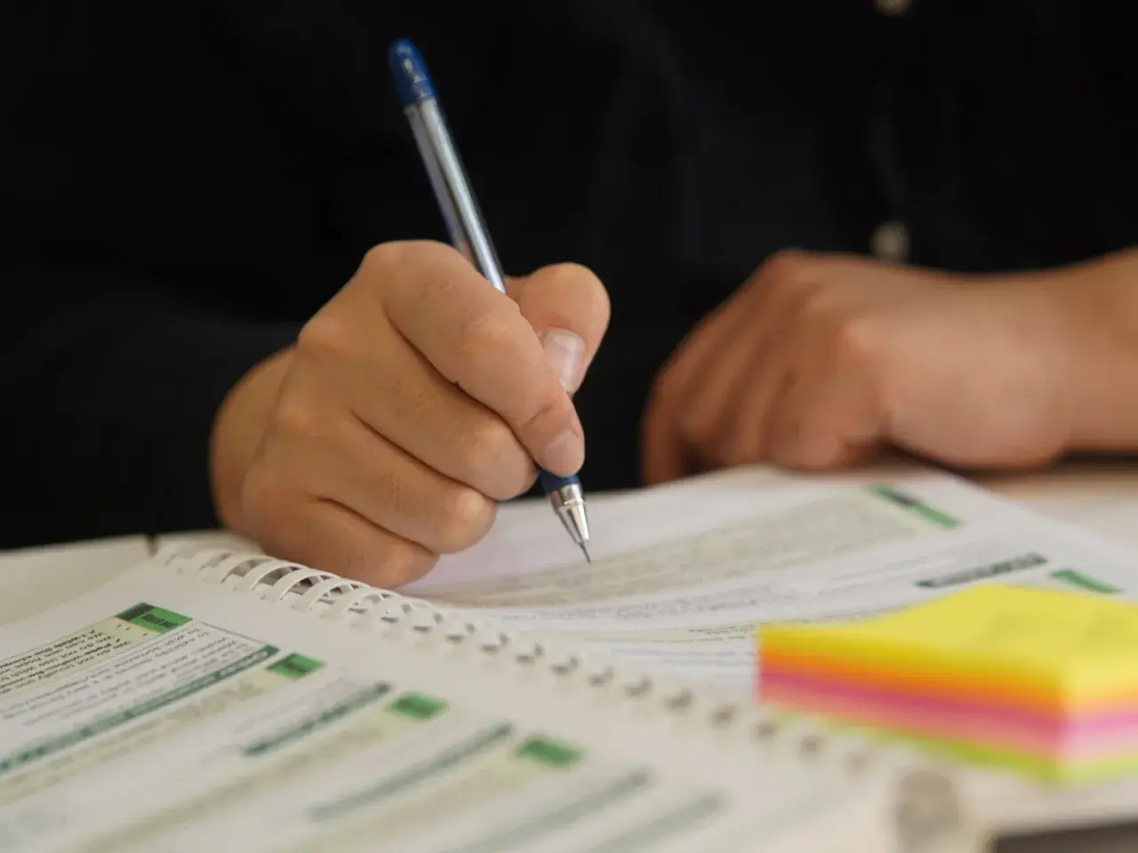 A professional engaging in language learning and translation, carefully reviewing a textbook and taking notes with a blue pen and colourful sticky notes.