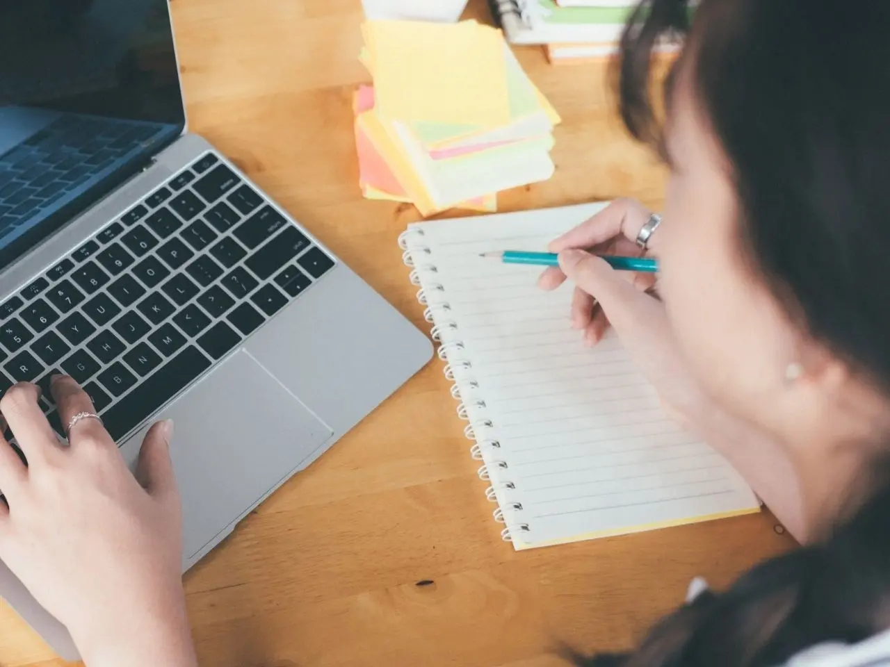 A person writes in a spiral notebook with a pencil while using a laptop on a wooden desk. Colourful sticky notes and other stationery are scattered nearby, suggesting a study or work in progress.