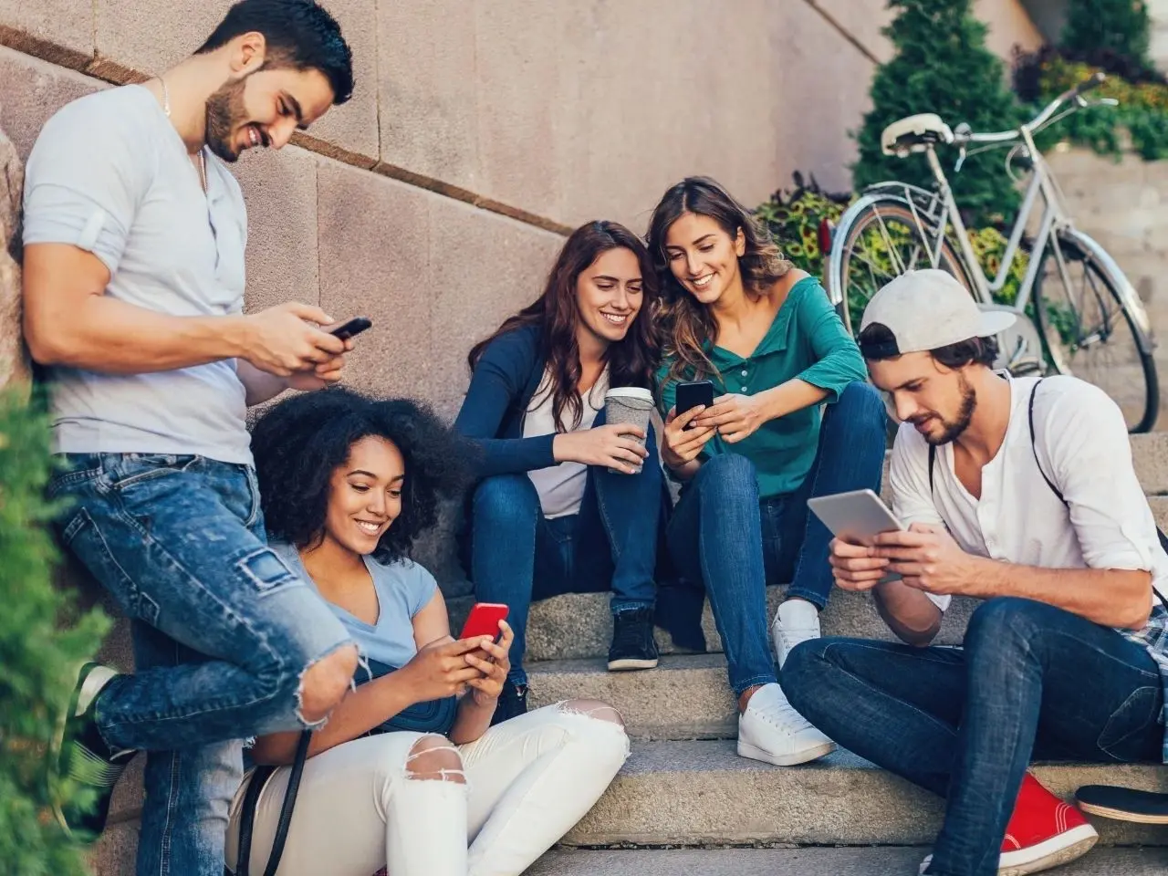 A group of five young adults sit and stand on outdoor steps, smiling and interacting with their smartphones and tablet. They appear relaxed and social, with a bicycle and greenery in the background.
