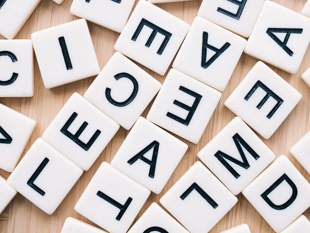 A scattered arrangement of white letter tiles with black uppercase letters, spread across a light wooden surface. The tiles appear random, with various letters oriented in different directions.