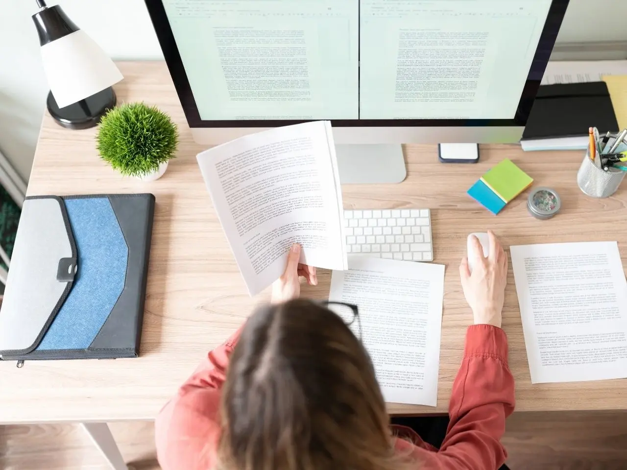 Overhead view of a woman working at a desk with documents and a large computer monitor. She is comparing printed text with digital content on a split-screen display. The desk is organized with a lamp, keyboard, notepad, pens, sticky notes, and a small potted plant.