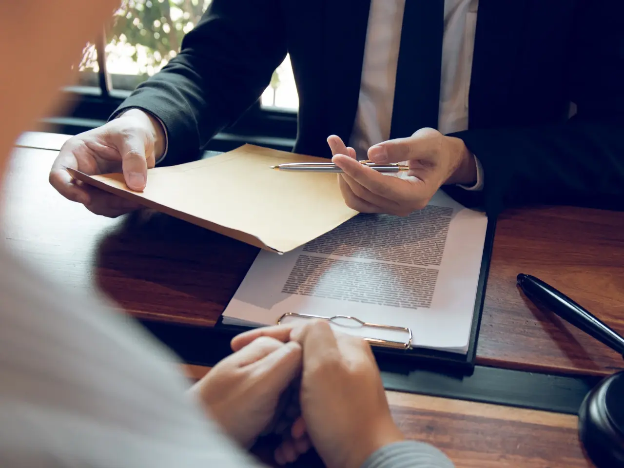 A professional setting where a person in a suit hands over a manila envelope and pen to another individual, suggesting the signing or exchange of legal documents. A clipboard with printed paperwork is visible on the wooden desk, indicating a formal or legal consultation involving a document apostille.