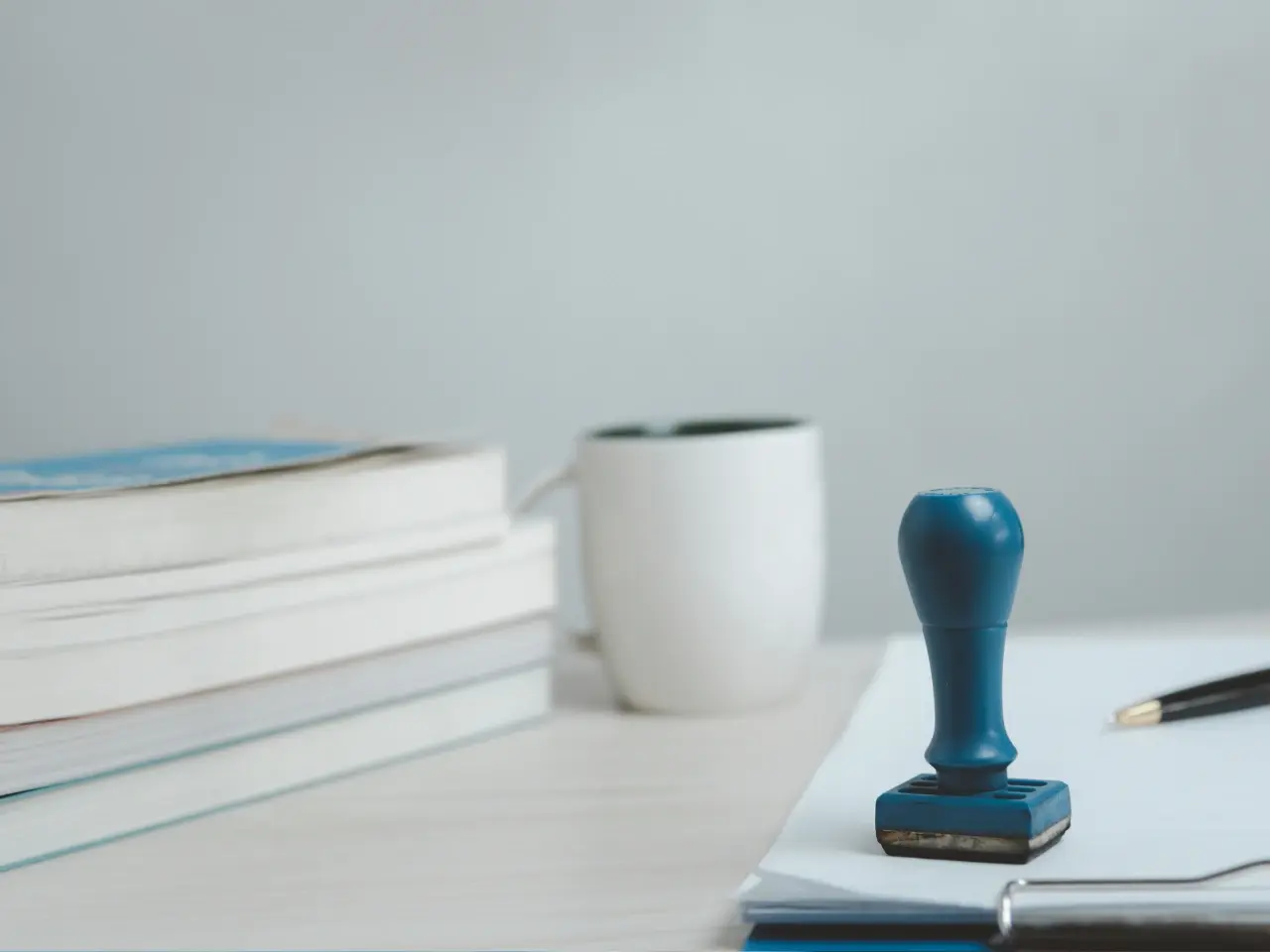 A close-up of a blue rubber stamp placed on top of a document on a desk. In the background, a white ceramic mug and a stack of books are slightly out of focus, along with a black pen and clipboard, suggesting an office or certification workspace.