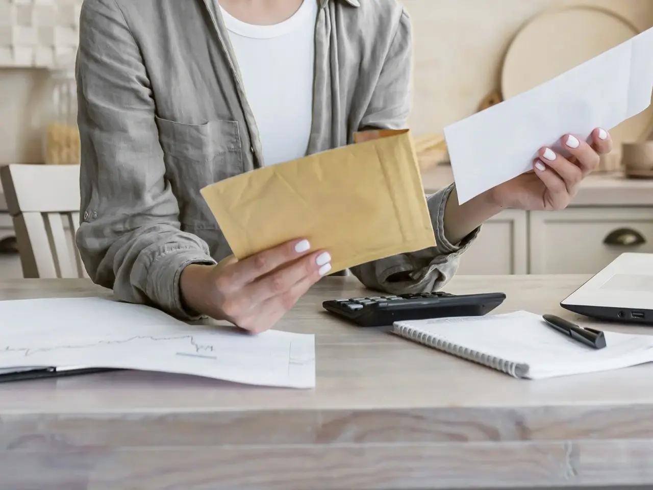 A woman seated at a table holds a padded yellow envelope in one hand and a white mailing envelope in the other. The workspace features documents, a notebook, a pen, a calculator, and a laptop, suggesting she’s preparing paperwork for mailing or organizing official correspondence.