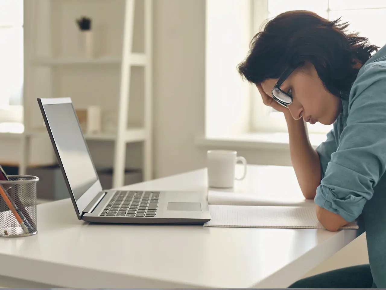A woman with dark hair and glasses sits at a desk in front of a laptop, resting her head on one hand and looking stressed or overwhelmed. A notebook, coffee mug, and pencil holder are also on the desk, suggesting she may be struggling with work or study-related tasks.