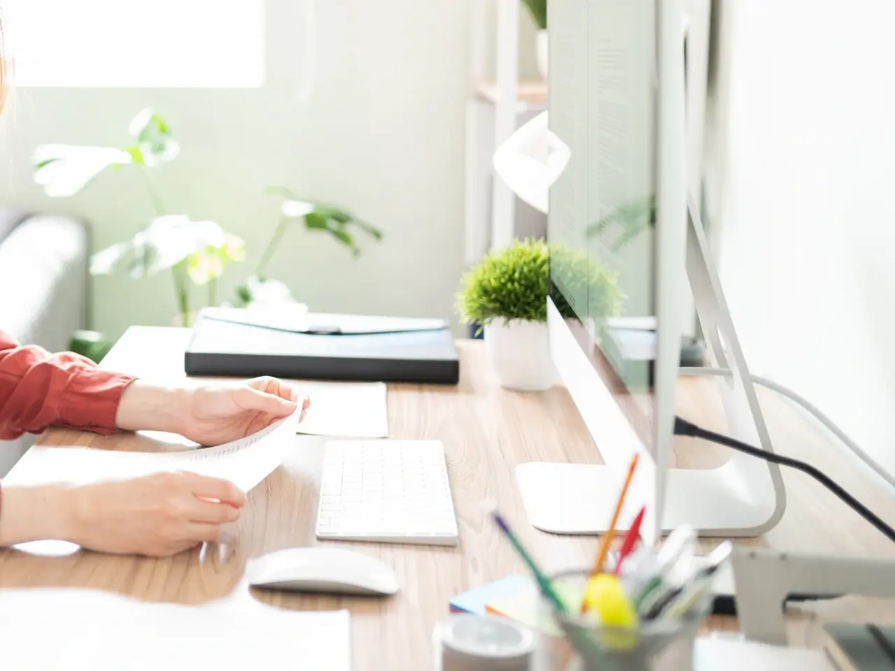 A well-lit home office with a modern desktop computer on a wooden desk. A person in a red shirt is reviewing documents, with a keyboard, mouse, potted plant, and office supplies nearby. Bright daylight and green plants add a fresh, productive atmosphere.
