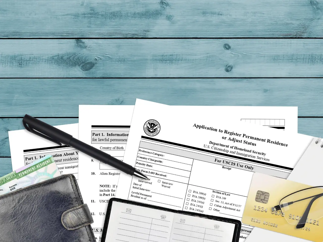 Application forms for permanent residence and adjustment of status on a desk with a pen, wallet, credit card, and glasses.