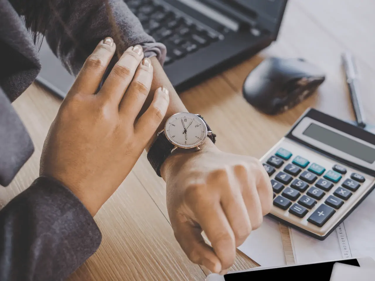 Person checking the time on a wristwatch at a desk with a calculator and laptop, symbolizing processing time for obtaining a Single Status Certificate in Canada.