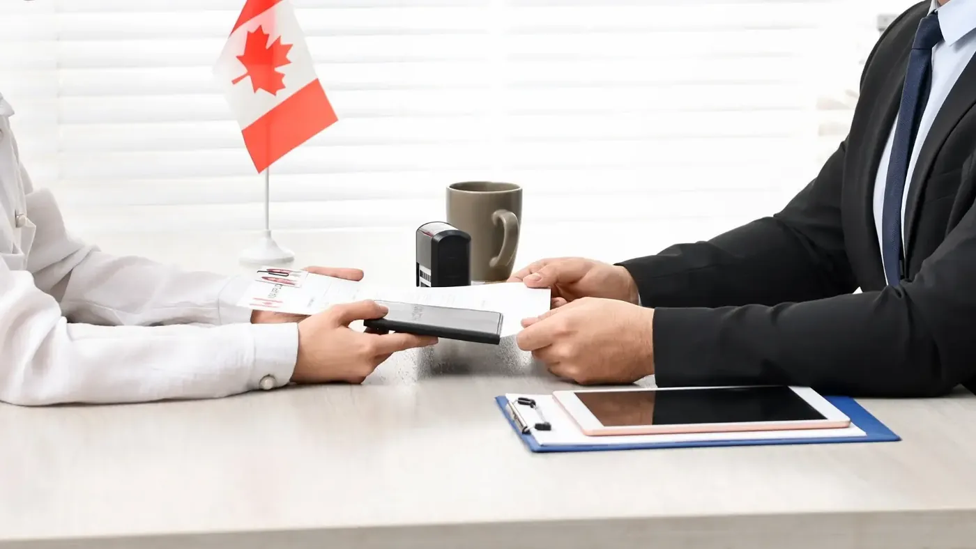 Two people at a table exchanging documents in a professional setting, with a Canadian flag, a stamp, a clipboard, and a tablet visible on the table, indicating an official or legal process in Canada.