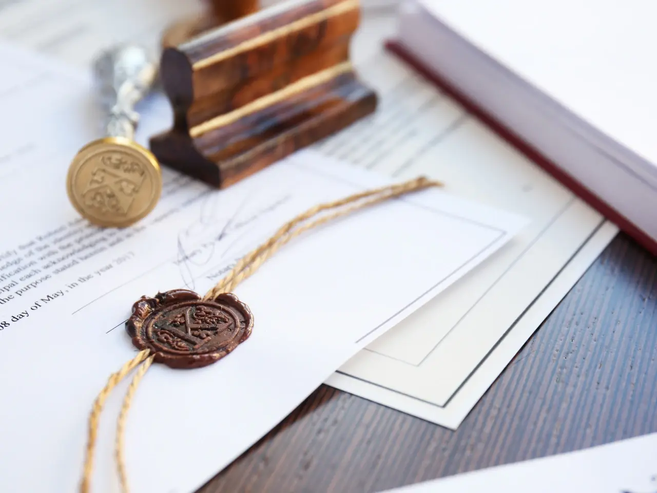 Close-up of a formal document with a red wax seal attached by a golden string, accompanied by a decorative metal stamp and a wooden seal press — evoking a sense of authenticity and tradition.