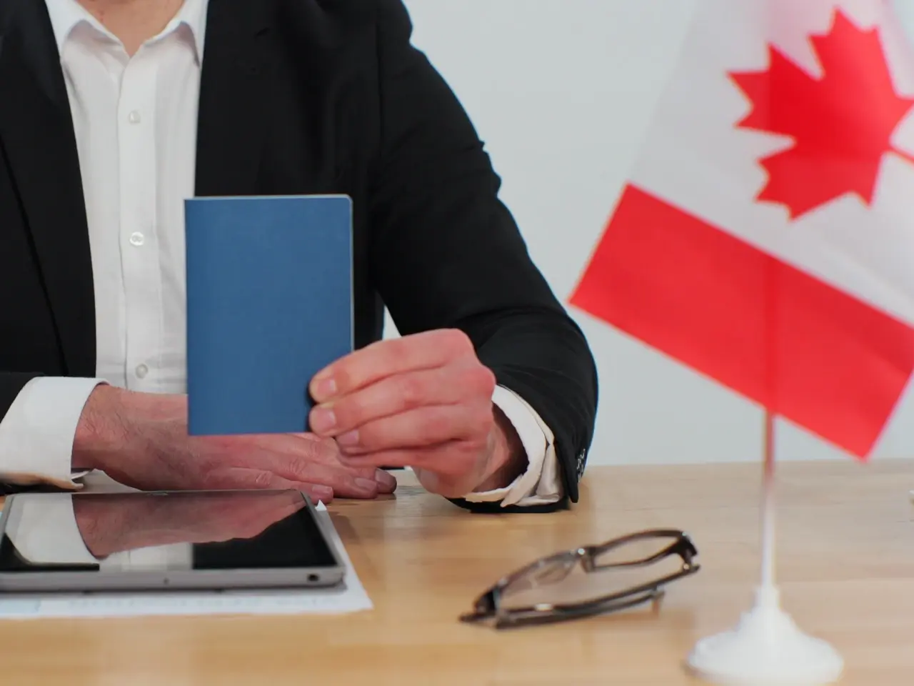 Person in a black suit holding a blue passport at a desk with a Canadian flag, tablet, and eyeglasses — suggesting an immigration, visa, or citizenship-related setting in Canada.
