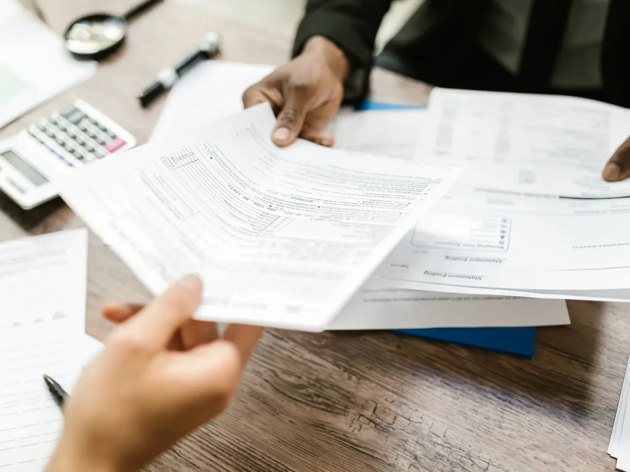 Close-up of hands exchanging filled-out government or financial forms over a wooden desk, with additional paperwork, a pen, calculator, and documents visible — suggesting a tax, legal, or immigration consultation.
