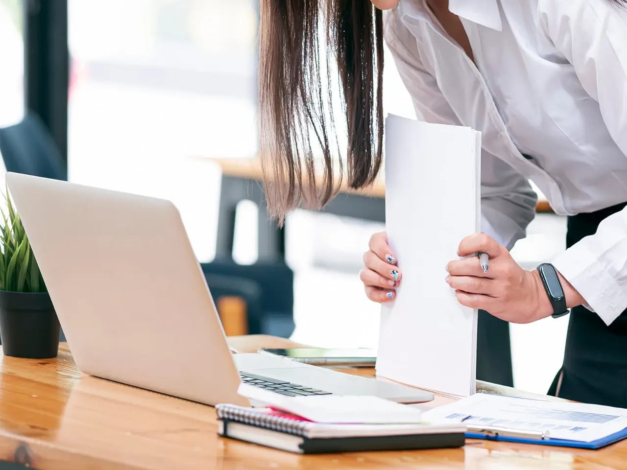 Professional woman in formal attire standing at a wooden desk, organizing documents beside an open laptop and notebooks, suggesting administrative work, translation, or client support tasks.