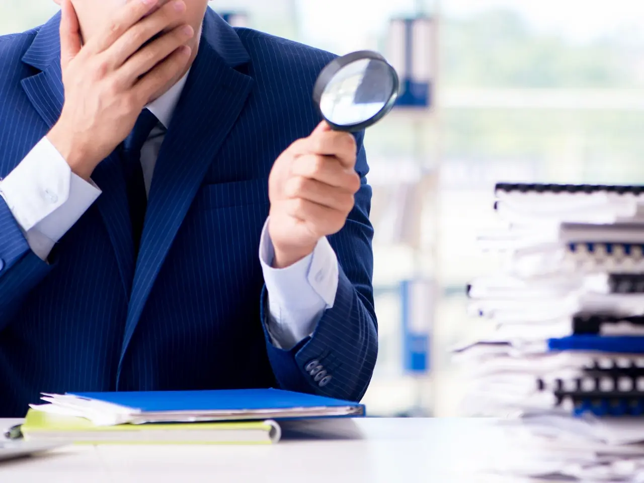 Businessman in a blue suit examining documents with a magnifying glass, surrounded by large stacks of paperwork, suggesting detailed review, legal scrutiny, or financial auditing.