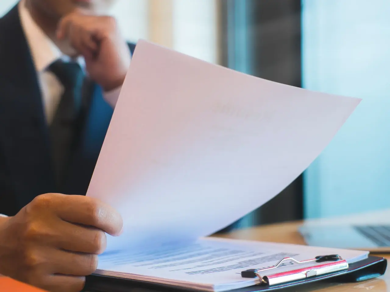 Close-up of a businessman in a suit reviewing a printed document on a clipboard, with a laptop nearby. The focus is on the paper, suggesting analysis, decision-making, or contract evaluation.