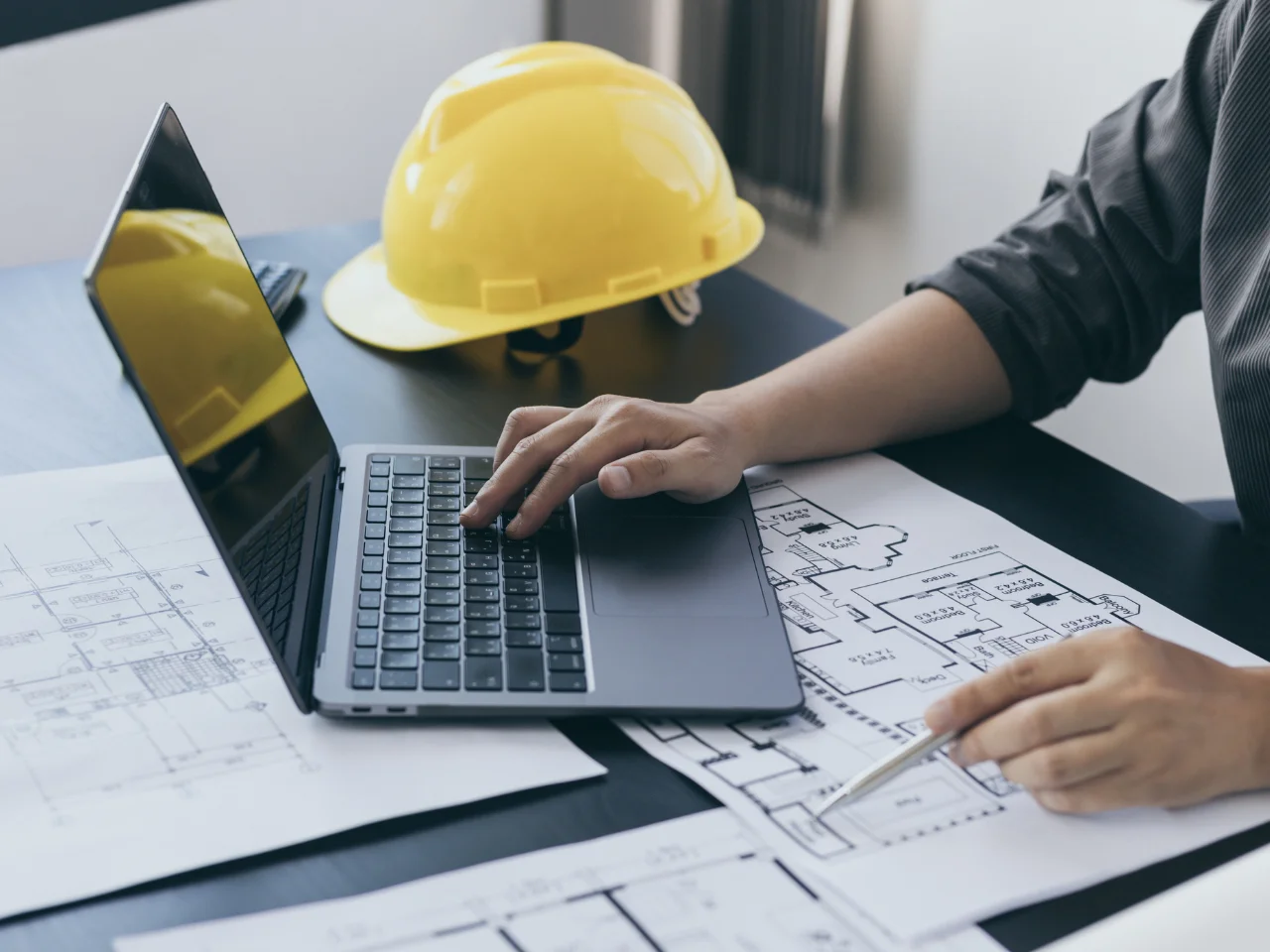 Professional engineer reviewing building plans on a laptop with a yellow hard hat on the desk.