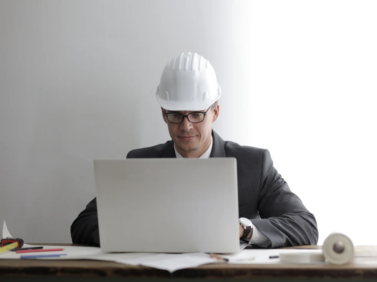 Engineer wearing a hard hat and reviewing plans on a laptop at a desk.